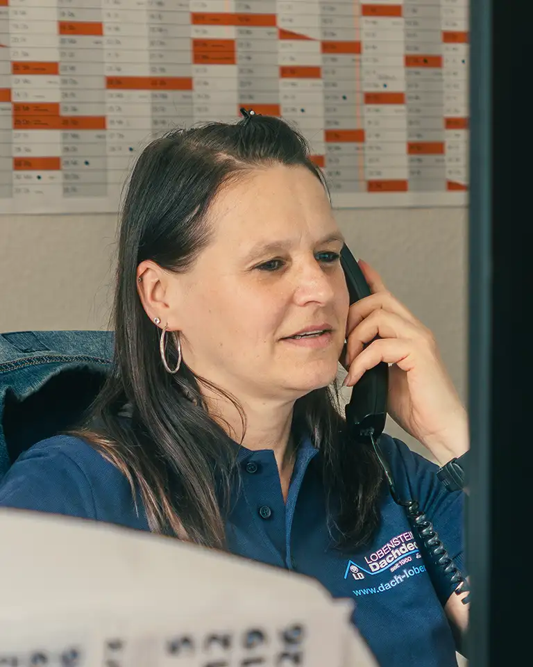 Woman in blue shirt talking on a corded phone at her desk with a calendar behind her.