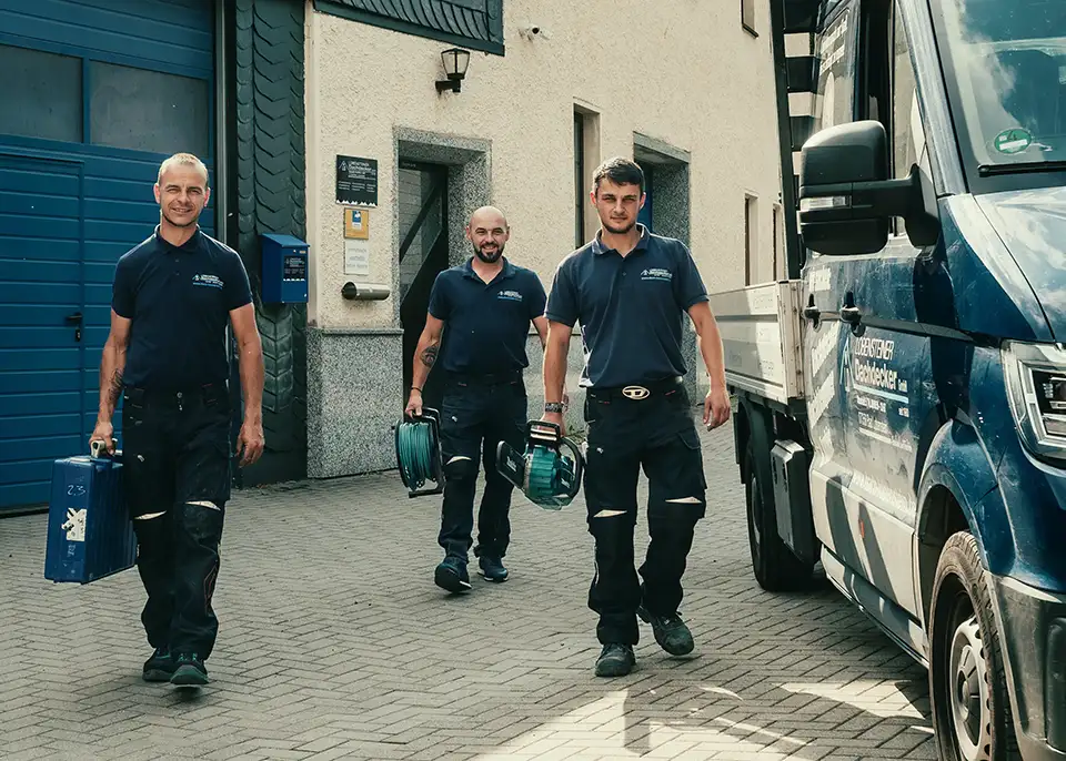 Three workers in navy uniforms carrying tools walking on paved ground near a building and a blue truck.
