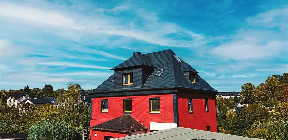Large white modern house with multiple dormer windows surrounded by green bushes and trees.