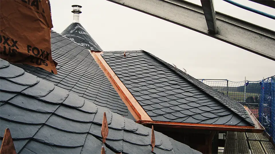 Wood-framed window set in dark slate-tiled roof with copper edging on a building under scaffolding.