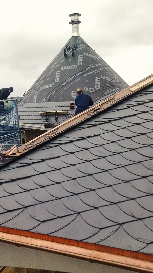 Wood-framed window set in dark slate-tiled roof with copper edging on a building under scaffolding.