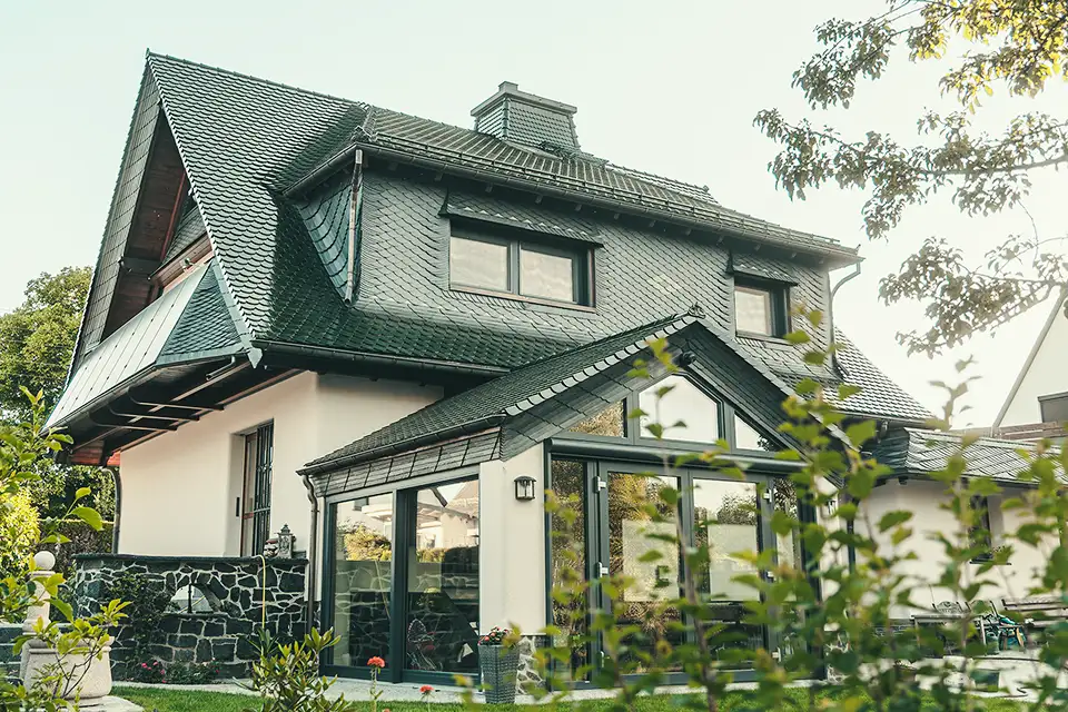 Modern house with dark tiled roof, large glass sunroom, surrounded by greenery.