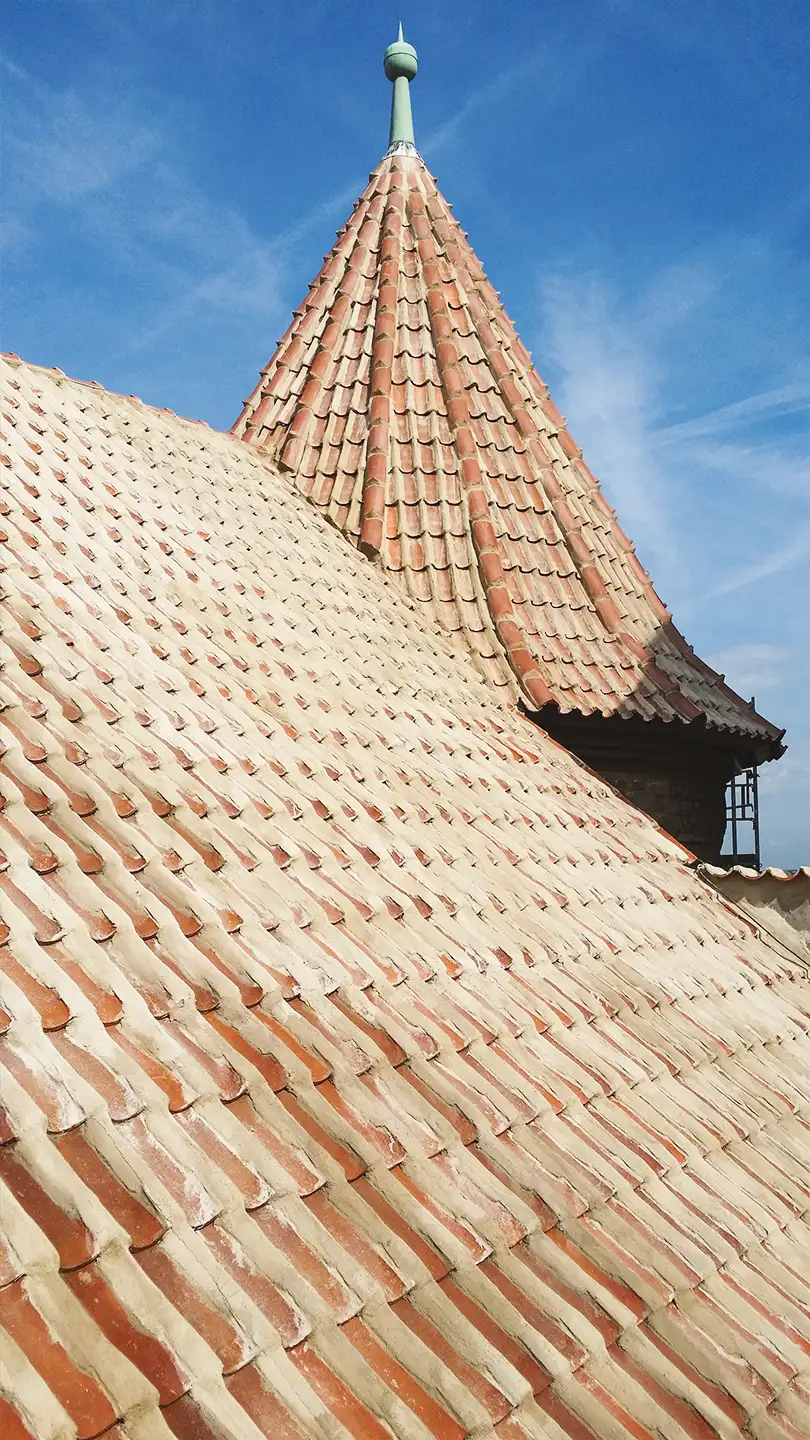 Curved dark slate roof with an attached rusty vertical pipe or antenna against a partly cloudy sky.