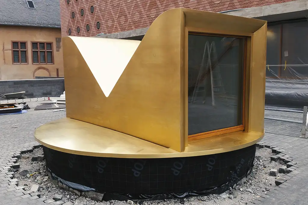 Modern gold-colored outdoor structure surrounding a window in a construction site.
