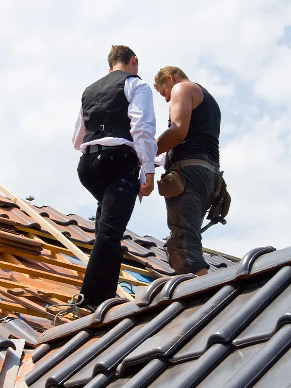 Two men standing on a tiled roof, one in work gear and the other in a vest and shirt under a cloudy sky.