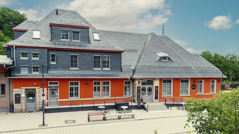 A historic railway station building with a gray slate roof and bright orange lower walls under a partly cloudy sky.