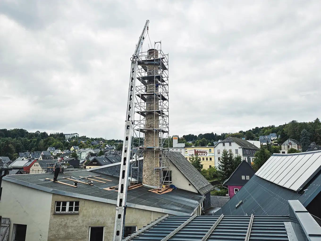 Man standing on flat rooftop near tall chimney scaffolded for repair in a town with cloudy sky.