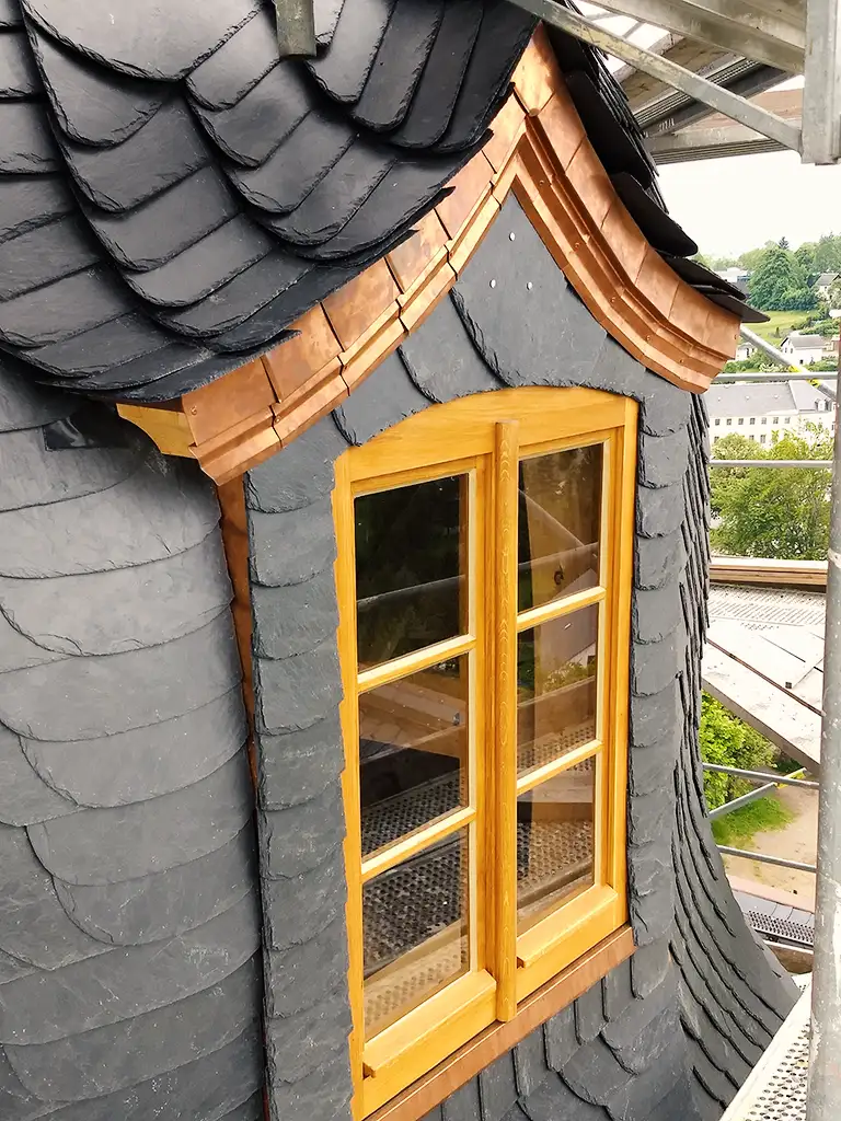 Wood-framed window set in dark slate-tiled roof with copper edging on a building under scaffolding.