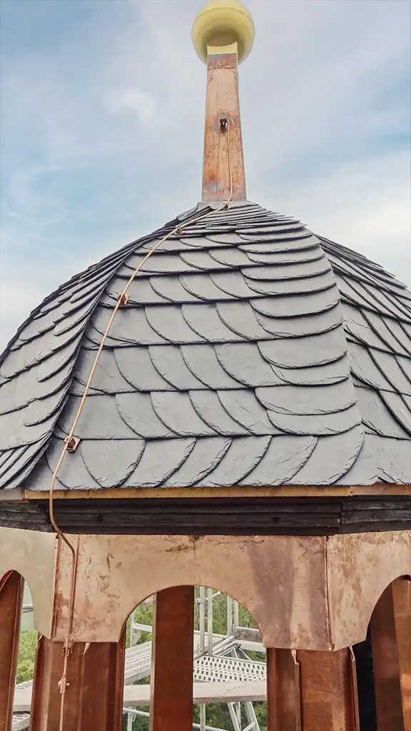 Curved dark slate roof with an attached rusty vertical pipe or antenna against a partly cloudy sky.