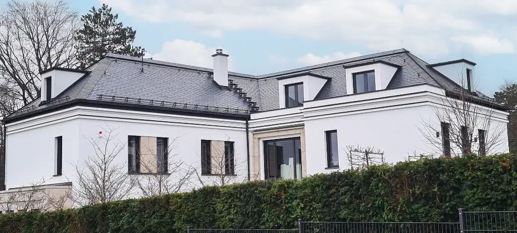 Large white modern house with multiple dormer windows surrounded by green bushes and trees.